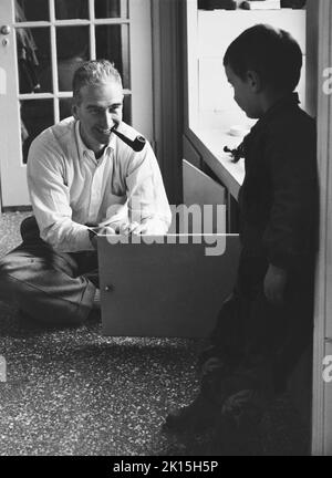 Ein Pfeifenraucher Vater öffnet eine Schranktür, während sein Sohn zuschaut. Ca. 1960. Stockfoto