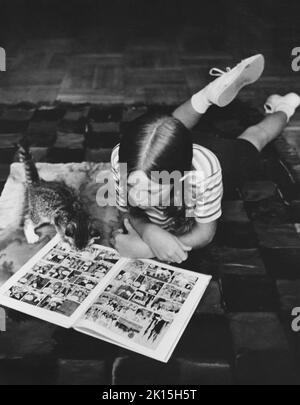 Ein Mädchen und ihr Kätzchen lagen zusammen auf einem Teppich und lasen ein Buch mit Cartoons. Ca. 1960, 1970. Stockfoto