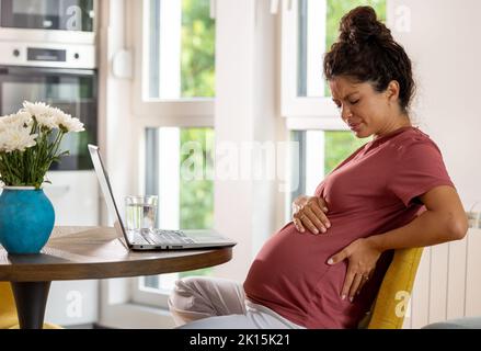 Eine Schwangerin sitzt auf einem Stuhl im Tisch mit einem Laptop vor ihr und hat Rückenschmerzen nach Überlastung Stockfoto