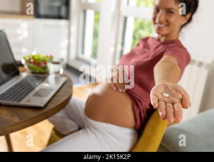 Nahaufnahme der weiblichen Hand mit Pille in der Handfläche einer Schwangeren, die am Tisch und Laptop sitzt Stockfoto