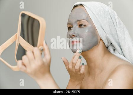 Mädchen legt graue kosmetische Tonmaske auf ihr Gesicht, während sie in Spiegel schaut, ihre Haare und ihr Körper sind in ein Handtuch gewickelt. Nahaufnahme Stockfoto