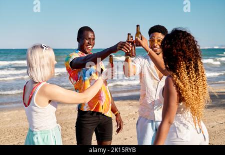 Fröhliche junge, vielfältige und beste Freunde, die am sonnigen Sommertag mit Flaschen Bier vor wolkenlosem, blauem Himmel lächeln und toasten Stockfoto