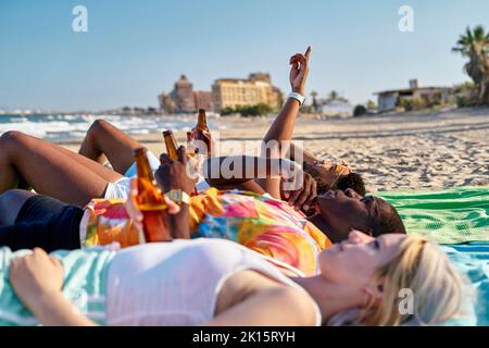 Seitenansicht von jungen multiethnischen männlichen und weiblichen Freunden in legerer Kleidung, die Bier trinken und auf den wolkenlosen blauen Himmel zeigen, während sie am Sandstrand liegen Stockfoto