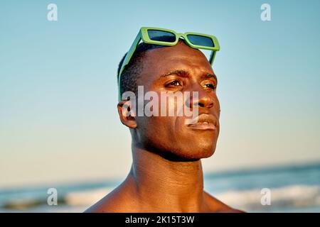 Seriöser afroamerikanischer Mann mit grüner Sonnenbrille auf dem Kopf, der auf das Meer und den wolkenlosen Sonnenuntergang blickt Stockfoto