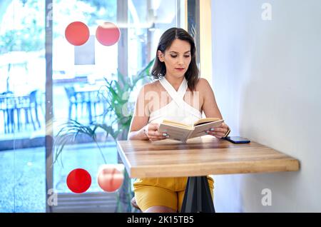 Aufmerksame junge Frau liest interessante Geschichte in einem Buch, während sie am Tisch sitzt und das Wochenende im Café verbringt Stockfoto