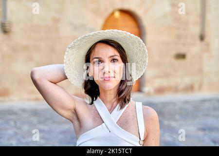 Porträt einer lächelnden erwachsenen Touristenattraktion mit Sonnenhut und Oberteil, die im Sommer vor dem verschwommenen Hintergrund der Altstadt die Kamera anschaut Stockfoto