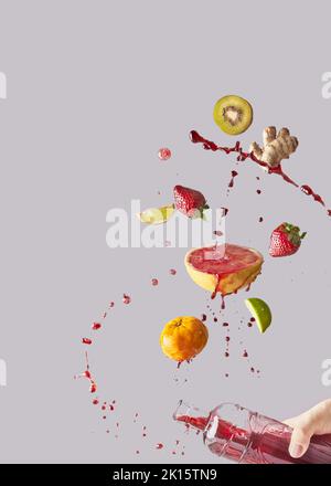 Ein Haufen aus verschiedenen frischen Früchten und Ingwer, der in eine Glasflasche fällt, wird von einer anonymen Person mit gesundem Saft vor grauem Hintergrund gehalten Stockfoto