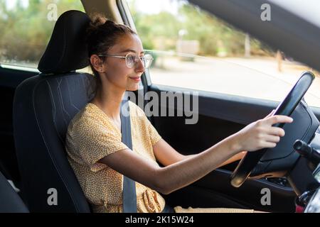 Junge Frau in legerer Kleidung und Brille, die wegschaut und an sonnigen Tagen ein modernes Fahrzeug fährt Stockfoto
