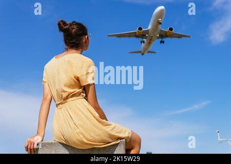 Von unten Rückansicht einer nicht erkennbaren Frau in legerer Kleidung, die am blauen Himmel sitzt und das Flugzeug am blauen Himmel fliegt Stockfoto
