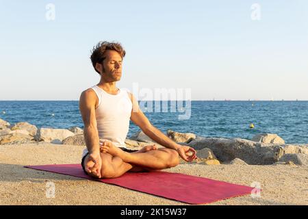 Barfuß-Mann in aktiver Trainingsbekleidung sitzt auf der Matte in Padmasana-Pose und meditiert mit geschlossenen Augen während der Yoga-Sitzung am Strand in der Nähe des Meeres am Morgen Stockfoto