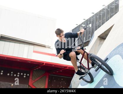 Junger Mann in legerer Kleidung, der am Wochenende auf dem BMX-Fahrrad auf einer Betonwand reitet und Zeit im Skatepark verbringt Stockfoto