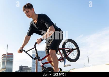 Von der unteren Seitenansicht des FOCUS-Mannes in schwarzem T-Shirt und Shorts in der Nähe einer Betonwand auf einem BMX-Fahrrad, während er auf der Stadtstraße Tricks macht Stockfoto