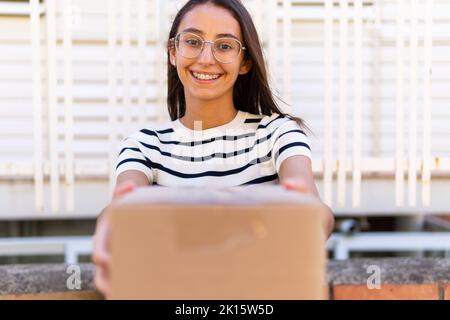 Entzückte junge gebärende Frau mit langen dunklen Haaren in gestreiftem T-Shirt und Brillen lächelnd und Blick auf die Kamera, während Pappkarton während o Stockfoto