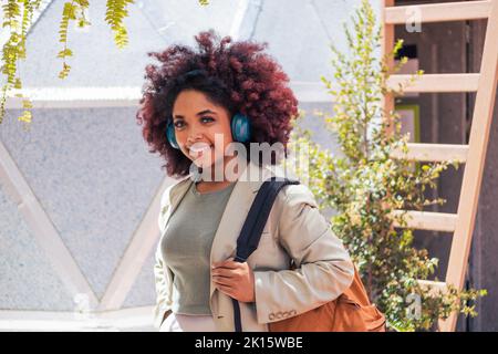 Optimistische junge afroamerikanische Managerin im Anzug mit Rucksack, die lächelnd auf die Kamera schaut und Musik hört, während sie gegen Geom steht Stockfoto
