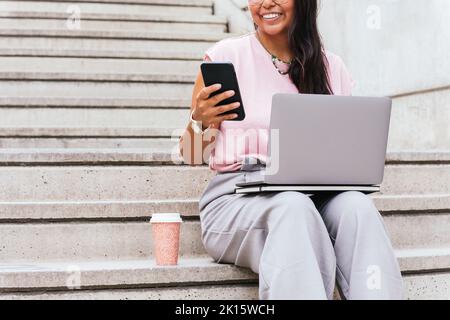 Crop anonyme ethnische Freiberuflerin mit dunklem Haar in elegantem Casual-Outfit und Brille lächelnd und mit Smartphone, während auf der Treppe mit sitzen Stockfoto