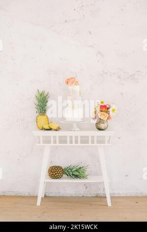 Frische Ananas und leckerer Kuchen, der während der Hochzeitsfeier neben den roten Schuhen mit hohen Absätzen an der weißen Wand auf den Tisch gelegt wird Stockfoto