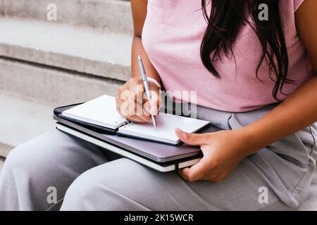 Crop anonyme Freiberuflerin mit langen dunklen Haaren in eleganten Freizeitkleidung sitzen auf Steintreppen mit Notebook und Laptop und Notizen im Plan Stockfoto