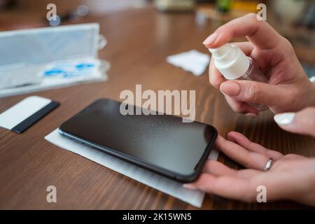 Weicher Fokus der anonymen Frau, die das Reinigungsprodukt auf dem Bildschirm des Mobiltelefons aufgibt, während sie das Schutzglas auf dem Tisch wechselt Stockfoto