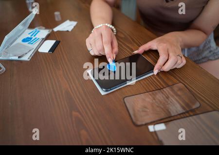 Von oben anonyme Frau mit Staubabsorber zu reinigen Bildschirm des Mobiltelefons, bevor Schutzglas auf dem Tisch kleben Stockfoto