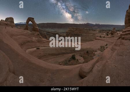 Atemberaubende Aussicht auf Delicate Arch in der Nähe des rauen Canyons vor der glühenden Milchstraße am Nachthimmel im Arches National Park in Utah, USA Stockfoto