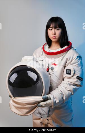 Junge asiatische Frau in Raumanzügen mit Helm und Blick auf die Kamera unter blauem Licht vor der Mission vor grauem Hintergrund Stockfoto