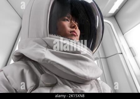 Asiatische Frau in Astronaut-Kostüm und Helm wegschauen während der Weltraummission im Raumschiff Stockfoto