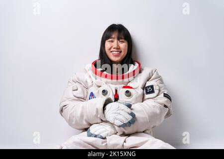 Junge asiatische Frau im Raumanzügen, die während der Weltraummission in der Nähe der grauen Wand sitzt und mit einem Lächeln auf die Kamera blickt Stockfoto