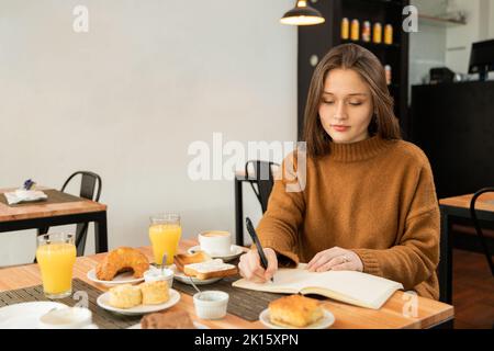 Junge Frau in braunem Pullover, die beim Frühstück mit Speisen und Getränken am Tisch sitzt und im Planer schreibt, während sie während des Frühstücks im Restaurant aus der Ferne arbeitet Stockfoto