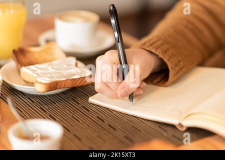 Eine nicht erkennbare Frau in braunem Pullover, die am Tisch sitzt und Frühstück und Getränke serviert und im Planer schreibt, während sie während des Bre aus der Ferne arbeitet Stockfoto