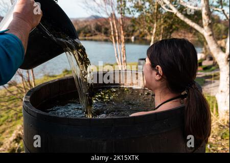 Crop männlichen Gießen Wasser mit Algen in Holzfass mit Brünette heißen Bad am Ufer des Sees im Herbst Stockfoto