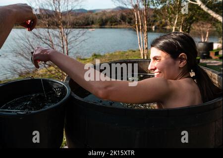 Ernte männlich Wasser mit Algen in Holzfass mit lächelnden Brünette unter heißen Bad am Ufer des Sees im Herbst Stockfoto