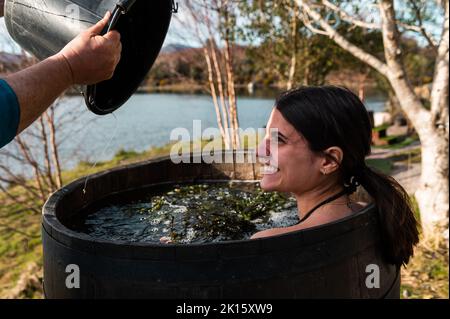 Ernte männlich Wasser mit Algen in Holzfass mit lächelnden Brünette unter heißen Bad am Ufer des Sees im Herbst Stockfoto