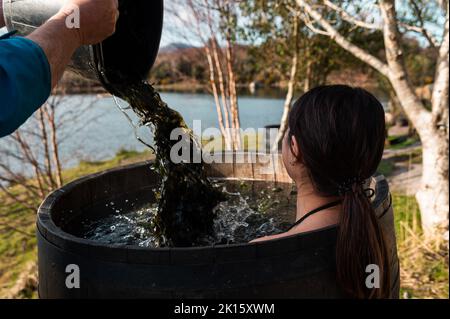 Crop männlichen Gießen Wasser mit Algen in Holzfass mit Brünette heißen Bad am Ufer des Sees im Herbst Stockfoto