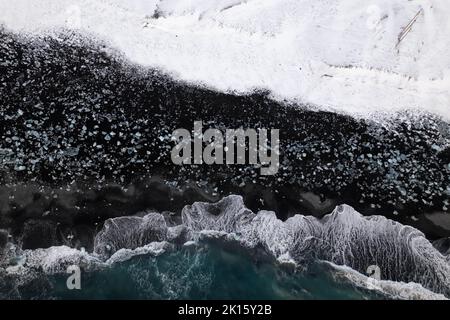 Luftaufnahme der schäumenden Meereswellen, die am kalten Wintertag in Island an der rauen, mit Schnee bedeckten Felsküste plantschen Stockfoto