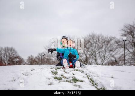 Zwei Kinder gleiten im Winter gemeinsam einen verschneiten Hügel hinunter Stockfoto