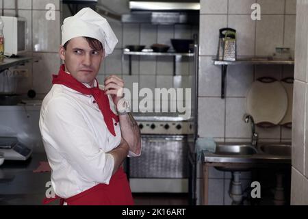 Junger gutaussehender Koch posiert in der Küche im Restaurant. Porträt eines positiven männlichen Köchers in Uniform Stockfoto