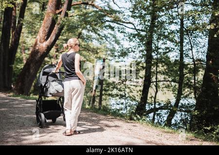 Rückansicht einer lässig gekleideten Mutter, die mit einem Kinderwagen im Stadtpark läuft. Konzept für Familie, Kind und Elternschaft. Stockfoto