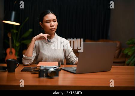 Fokussierte und professionelle, tausendjährige asiatische Geschäftsfrau oder Chefin, die nachts von zu Hause aus arbeitet und an ihrem Projekt mit Laptop-Computing arbeitet. Stockfoto