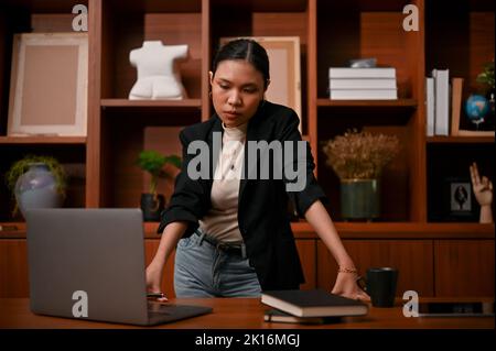 Professionelle und entschlossene, tausendjährige asiatische Geschäftsfrau oder weibliche Chefin steht im Büro, durchdacht und pensives Planen ihrer Business-Strat Stockfoto