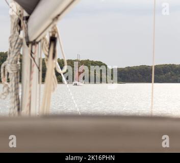 Ein ruhiger Blick auf ein kleines rotes Boot, das von einem größeren Schiff aus gesehen wird und auf einem ruhigen Fluss schwimmt, umgeben von üppigen grünen Wäldern unter einem bewölkten Himmel. Stockfoto