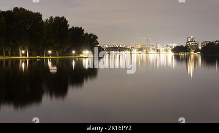 Die nächtliche Stadtlandschaft spiegelt sich in einem ruhigen See wider, mit leuchtenden Straßenlaternen und beleuchteten Gebäuden, die eine ruhige und malerische urbane Szene schaffen. Stockfoto