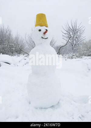 Ein Schneemann aus drei Schneebällen, er hat einen gelben Eimer auf dem Kopf, eine Karotte statt einer Nase und Kohlen statt Augen. Vor dem Hintergrund von Stockfoto