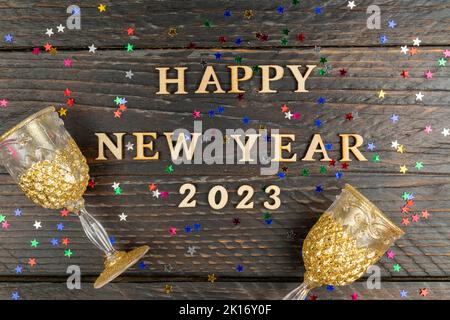 Holztext glückliches neues Jahr 2023 auf Holzhintergrund umrahmt bunten Sternen Konfetti und zwei Gläser für Champagner. Festliche Grußkarte. Stockfoto