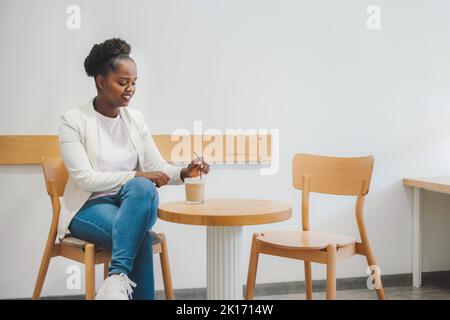 Eine verträumte junge afrikanische Geschäftsfrau am Couchtisch, die kaltes Getränk trinkt und nach unten schaut. Genießen Sie das Aroma in der Cafeteria des Cafés. Spaß haben Stockfoto