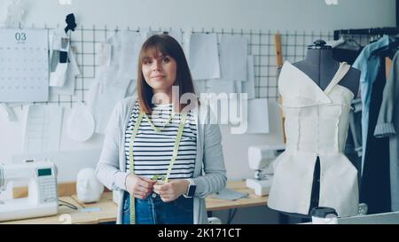 Porträt einer schönen jungen Näherin, die am Arbeitsplatz steht, das Maßband berührt und die Kamera anschaut. Dummy, Damenbekleidung, Skizzen und Nähmaschine sind sichtbar. Stockfoto