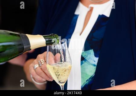 Nahaufnahme einer Champagnerflasche, die Champagner auf einer Sektflöte gegen eine verschwommene Person auf einer Party umschaufelt Stockfoto