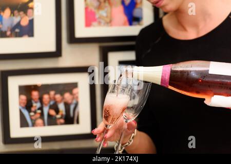 Nahaufnahme einer Champagnerflasche, die pinkfarbenen Champagner in eine Sektflöte vor einer verschwommenen Wand mit Fotos auf einer Party pochelt Stockfoto