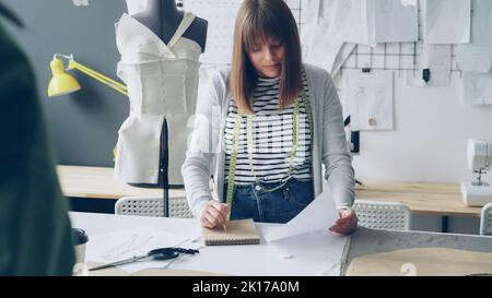 Der professionelle Schneidermeister macht Notizen in Notizblock und schaut sich die Kleidungsskizze an, während er an einem großen Studientisch arbeitet. Viele Zeichnungen, Dummy und Nähmaschine im Hintergrund. Stockfoto