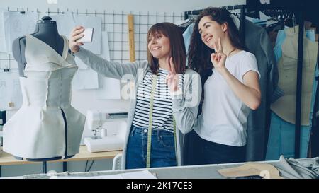 Zwei fröhliche weibliche Bekleidungsdesigner machen mit dem Smartphone ein lustiges Selfie, während sie neben einer gekleideten Schaufensterpuppe im Studio stehen. Attraktive Frauen lächeln und posieren. Stockfoto