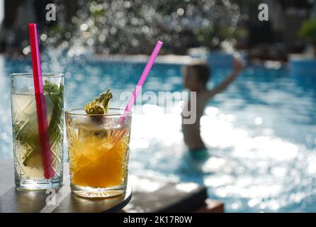 Konzentrieren Sie sich auf zwei Fruchtcocktails, die am Pool stehen, und das Kind spielt im Wasser im Hintergrund, perfekt für die Werbung Stockfoto